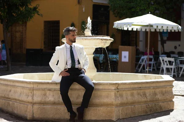 Handsome young man with beard and white jacket sitting on the edge of a fountain in a typical square in the city of seville in spain. The man is on holiday in the city