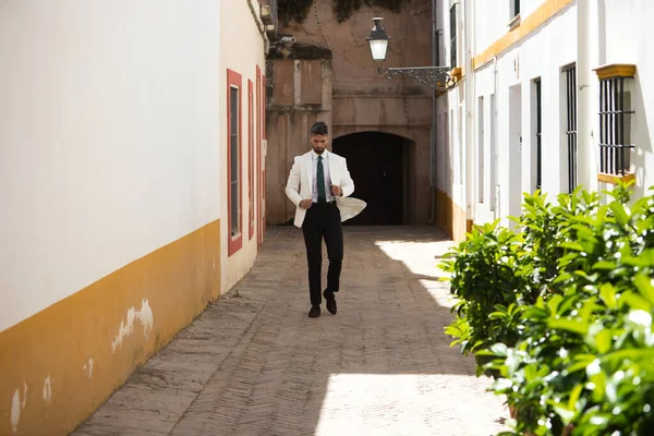 handsome young man with beard walking down the street unbuttons his white jacket. The man is on holiday in seville and walks through a typical neighbourhood of the city in spain. Holiday concept