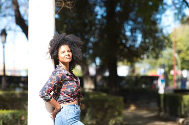 Parktaki mermer bir sütuna yaslanmış, afro saçlı, genç, güzel siyahi bir kadın. Kadın farklı ifadeler kullanırken güneşli bir günün tadını çıkarıyor.