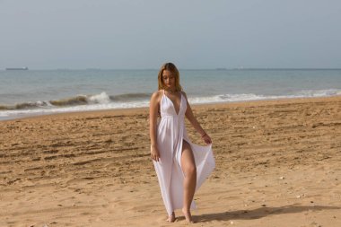 Beyaz elbiseli güzel sarışın kadın güneşli bir günde kumsalın kıyısında yürüyor. Kadın farklı vücut ifadeleri kullanıyor. Arka planda mavi deniz.