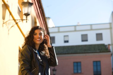 Young and beautiful Hispanic brunette woman with curly hair and black leather clothes is sitting on a railing in the street. The woman is happy and smiling.