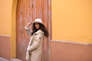 Young and beautiful Hispanic brunette woman with curly hair wearing a hat and coat for the cold walking in the city of seville while making different expressions and having fun.