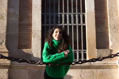 young beautiful brunette woman with curly hair and green woollen coat is leaning on the chains surrounding the cathedral of seville in spain. The woman is happy and enjoying her holiday in the city.