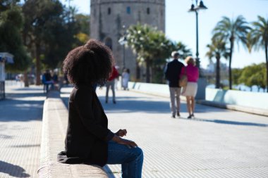 Genç, güzel, siyah saçlı bir kadın sokağın kenarına bakan bir bankta oturuyor. Seyahat ve tatil konsepti.