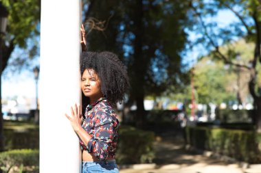 Parktaki mermer bir sütuna yaslanmış, afro saçlı, genç, güzel siyahi bir kadın. Kadın farklı ifadeler kullanırken güneşli bir günün tadını çıkarıyor.