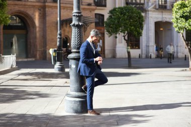 Sakallı, mavi takım elbiseli ve kravatlı, lambaya yaslanmış ve saate bakan yakışıklı genç adam, İspanya 'da Seville' de bir Avrupa şehrine iş gezisinde.