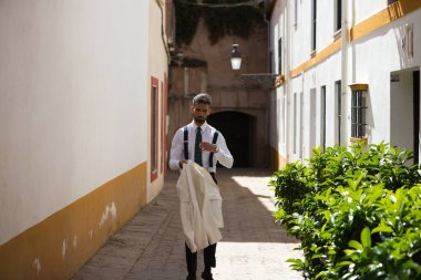 Sokakta yürüyen sakallı yakışıklı genç adam beyaz ceketini çıkarıyor. Adam Seville 'de tatilde ve İspanya' nın tipik bir mahallesinde yürüyor. Tatil kavramı.
