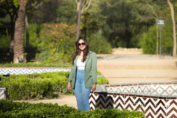 Молодая красивая испанская женщина с длинными каштановыми волосами в джинсах и зеленой куртке и солнечных очках гуляет по парку в Севилье, Испания. Женщина счастлива и улыбается на фото в свой праздник