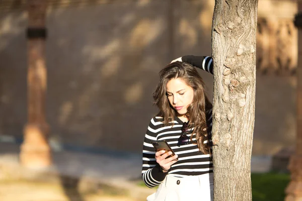 Uzun kahverengi saçlı, siyah beyaz çizgili tişört giyen, ağaca yaslanmış ve cep telefonuna bakan genç, güzel bir kadın. Yeni teknoloji ve telefon kavramı