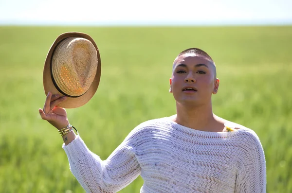 Portrait of a young non-binary latin person holding a straw hat in his hand and standing in the green wheat field making different expressions. Concept of diversity, homosexuality and human rights