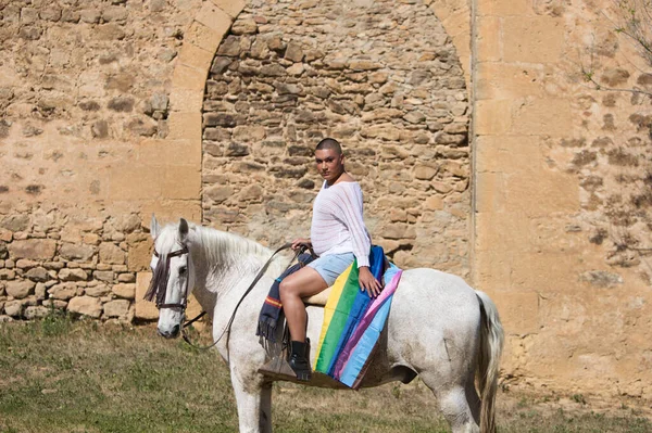 Young non-binary latin person, riding a white horse with the gay pride flag. In the background a medieval castle. Concept of diversity, homosexuality and human rights