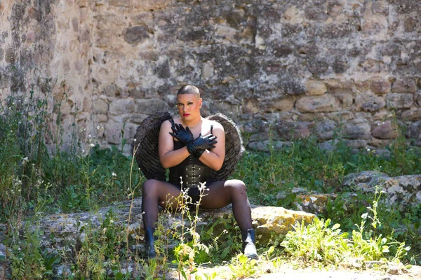 Young non-binary Latin person, wearing black make-up and black angel wings, sitting on a rock and covering his chest with his hands. Concept of diversity, homosexuality and human rights
