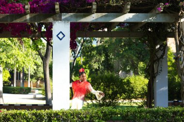 Flamenko elbiseli genç siyahi kadın İspanya 'nın Sevilla kentindeki İspanyol Plaza' sının yanındaki ünlü bir parkta begonvillerle birlikte bir sütuna yaslanıyor. Fırfırlı ve kırmızı şallı bej bir elbise giyiyor..