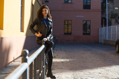 Young and beautiful Hispanic brunette woman with curly hair and black leather clothes is sitting on a railing in the street. The woman is happy and smiling.