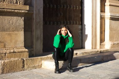 Kıvırcık saçlı, yeşil yün paltolu güzel esmer kadın Seville, İspanya 'da bir binanın girişinde oturuyor. Kadın mutlu ve şehirde tatilinin tadını çıkarıyor..
