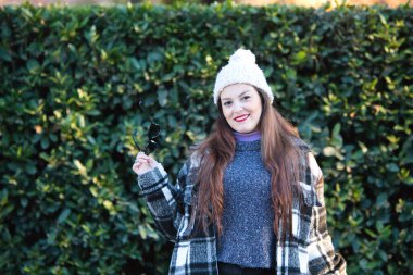 Ekose ceketli, yün şapkalı ve güneş gözlüklü güzel bir kadın parkta güneşli kış gününün tadını çıkarıyor. Kadın mutlu ve gülümsüyor..