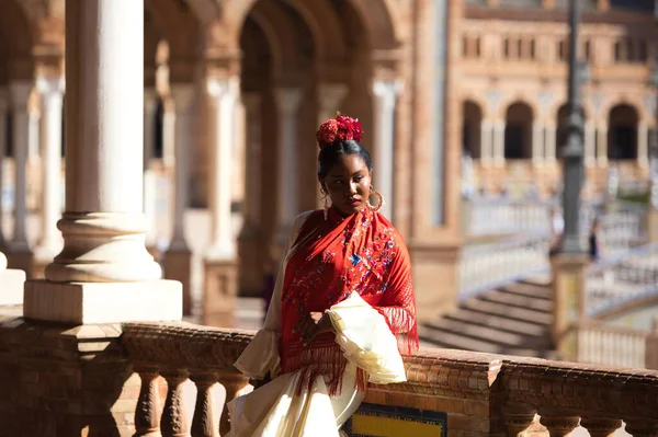 stock image Young black woman dressed as a flamenco gypsy in a famous square in Seville, Spain. She wears a beige dress with ruffles and a red shawl with flowers. Flamenco cultural heritage of humanity