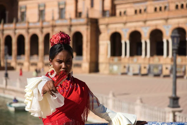 Young black woman dressed as a flamenco gypsy in a famous square in Seville, Spain. She wears a beige dress with ruffles and red shawl and red fan. Flamenco cultural heritage of humanity