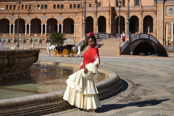 Young black woman dressed as a flamenco gypsy in a famous square in Seville, Spain. She is wearing a beige dress with ruffles and a red shawl and is standing in front of a fountain