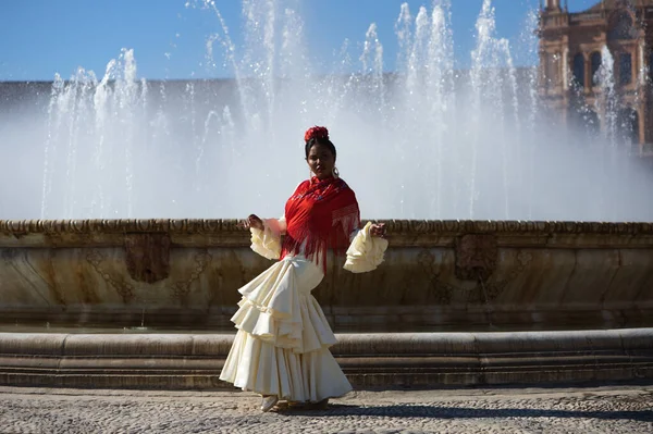 Young black woman dressed as a flamenco gypsy in a famous square in Seville, Spain. She is wearing a beige dress with ruffles and a red shawl and is standing in front of a fountain