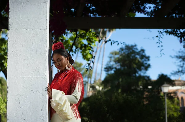 stock image Young black woman in flamenco dress leaning on a column with bougainvilleas in a famous park next to the Plaza de spain in Seville, Spain. She wears a beige dress with ruffles and red shawl