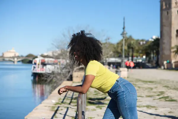 Young and beautiful black woman with afro hair and sunglasses wearing jeans and yellow shirt leaning on the railing overlooking the river in seville.