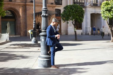 Sakallı, mavi takım elbiseli ve kravatlı, lambaya yaslanmış ve saate bakan yakışıklı genç adam, İspanya 'da Seville' de bir Avrupa şehrine iş gezisinde.