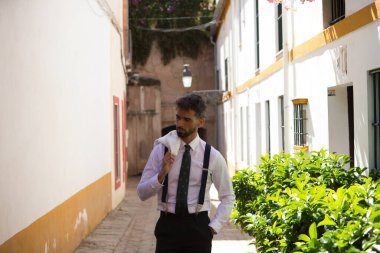 Sokakta yürüyen sakallı, omuzunda beyaz ceketi olan yakışıklı bir genç. Adam Seville 'de tatilde ve İspanya' nın tipik bir mahallesinde yürüyor.