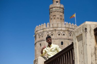 Sakallı ve elinde güneş gözlüğü olan yakışıklı genç adam korkuluklara yaslanıyor. Arka planda Seville 'deki nehrin kıyısındaki Torre del Oro anıtı var. Seyahat ve tatil konsepti.