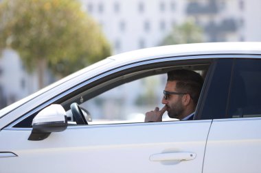 Beyaz spor arabasında oturan sakallı ve mavi takım elbiseli yakışıklı genç bir iş adamı. Adam güneş gözlüğü takıyor. İş seyahati kavramı