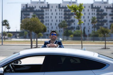 Sakallı ve mavi takım elbiseli yakışıklı genç iş adamı beyaz spor arabasına yaslanıyor. Adam güneş gözlüğü takıyor. İş seyahati kavramı