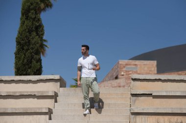 Günlük kıyafetler ve güneş gözlüğü takmış yakışıklı genç adam tribünün merdivenlerinden aşağı iniyor ve mekanın farklı yerlerine bakıyor.