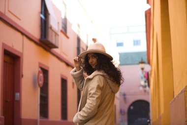 Young and beautiful Hispanic brunette woman with curly hair wearing a hat and coat for the cold walking in the city of seville while making different expressions and having fun.