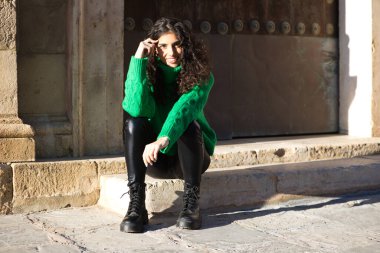 Kıvırcık saçlı, yeşil yün paltolu güzel esmer kadın Seville, İspanya 'da bir binanın girişinde oturuyor. Kadın mutlu ve şehirde tatilinin tadını çıkarıyor..