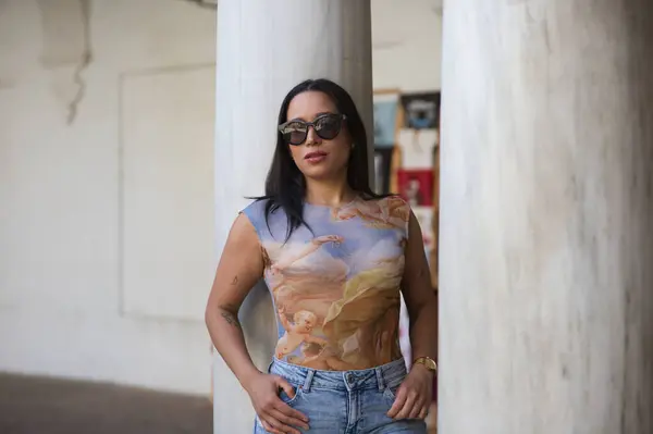 stock image Latin brunette woman with long hair and sunglasses is dressed in jeans and printed top. The girl is leaning between two marble columns. Travel and holiday concept