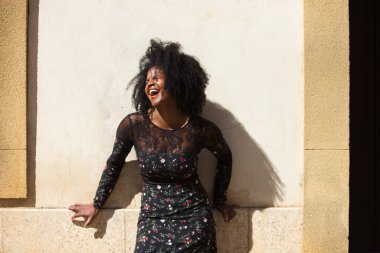 Siyah kadın portresi, genç ve güzel, afro saçlı ve çiçekli siyah elbiseli çok mutlu ve gülümsüyor. Arka planda açık kahverengi bir duvar var. Farklı ifadeleri kavra