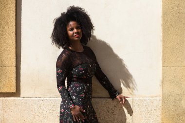 Siyah kadın portresi, genç ve güzel, afro saçlı ve çiçekli siyah elbiseli çok mutlu ve gülümsüyor. Arka planda açık kahverengi bir duvar var. Farklı ifadeleri kavra