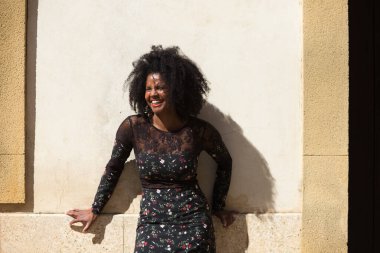 Siyah kadın portresi, genç ve güzel, afro saçlı ve çiçekli siyah elbiseli çok mutlu ve gülümsüyor. Arka planda açık kahverengi bir duvar var. Farklı ifadeleri kavra