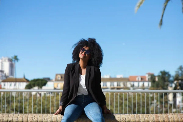 Young beautiful black woman with afro hair sitting on a bench wearing jeans and black jacket enjoying her holidays in spain. Travel and holiday concept.