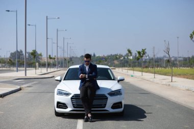 Sakallı ve mavi takım elbiseli yakışıklı genç bir işadamı beyaz spor arabasında kollarını kavuşturmuş saate bakıyor. Adam güneş gözlüğü takıyor. İş seyahati kavramı