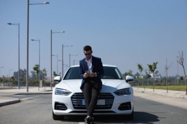 Sakallı ve mavi takım elbiseli yakışıklı genç işadamı beyaz spor arabasının yanında duruyor. Adam güneş gözlüğü takıyor ve cep telefonuna bakıyor. İş adamı ve seyahat konsepti.