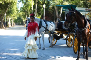 İspanya, Seville 'de ünlü bir meydanda flamenko çingenesi gibi giyinmiş genç siyahi bir kadın. Bej bir elbise ve fırfırlı kırmızı bir şal giyiyor ve at arabasının yanında duruyor.