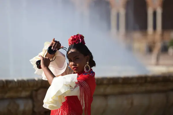 Young black woman dressed as a flamenco gypsy in a famous square in Seville, Spain. She is wearing a beige dress with ruffles and red shawl and playing castanets.Flamenco cultural heritage of humanity