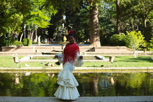 Young black woman dressed as a flamenco dancer stands in a famous park next to a duck pond in Seville, Spain. She wears a beige dress with ruffles and red shawl and flowers in her hair