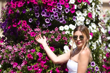 Sarı saçlı ve güneş gözlüklü güzel esmer kadın farklı renkte çiçeklerle dikey bir bahçenin önünde. Kadın farklı ifadeler kullanıyor.