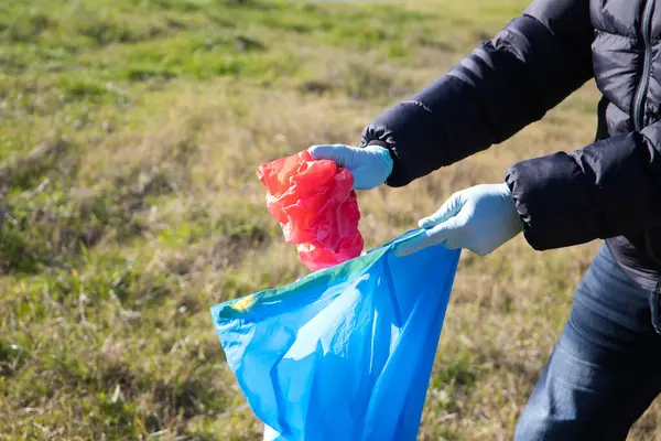 Detail of a volunteer\'s hand picking up a red plastic bag from the forest and putting it in a garbage bag. Concept of Earth Day and World Environment Day 5 June.
