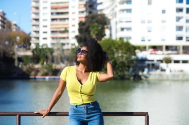 Genç, güzel, siyah saçlı, güneş gözlüklü kot pantolon giyen ve sarı gömlekli bir kadın korkuluklara yaslanıyor. Arka planda Guadalquivir nehri ve Sevilla mahallesi var..