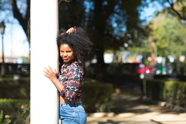 Young, beautiful black woman with afro hair dressed in casual clothes leaning against a marble column in the park. The woman enjoys the sunny day while making different expressions