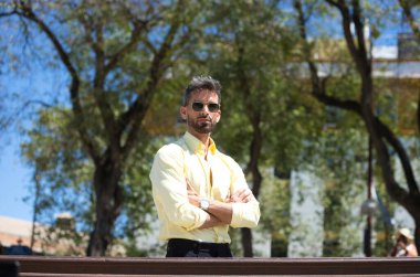 Sakallı, güneş gözlüklü, kolları katlanmış ve kendinden emin yakışıklı genç bir adam. Adam Seville 'de arka planda ağaçlarla bir bahçede.