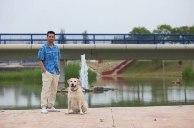 Sakallı ve Labrador av köpeği olan yakışıklı genç bir adam. Parktaki gölde fotoğraf çekmek için poz veriyor. Adam evcil hayvanına sevgisini okşayarak, öpücüklerle ve sarılmalarla gösterir. Evcil hayvanlar.